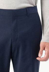 Hombre vistiendo pantalones ajustados azul marino y camisa de manga larga gris claro, con la mano derecha relajada a un lado.