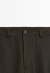 Close-up of dark brown pants waistband with belt loops and a single black button fastening on a white background.