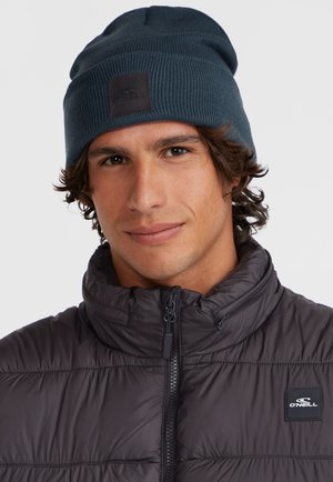 Dark blue knitted beanie with a textured finish and a black logo patch. Worn with a black puffer jacket featuring a high collar.