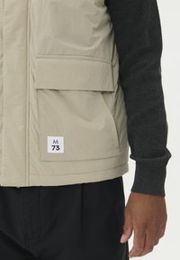 Bež prsluk s velikim prednjim džepovima i oznakom "M 73" nošen preko tamno sive dugih rukava i crnih hlača, ruka opuštena uz tijelo.