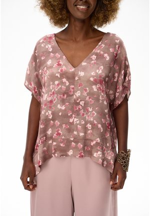 Kobieta ubrana w luźną różową kwiecistą bluzkę z dekoltem w kształcie litery V oraz jasnoróżowe spodnie, uzupełniona dużą złotą bransoletką na prawym nadgarstku.