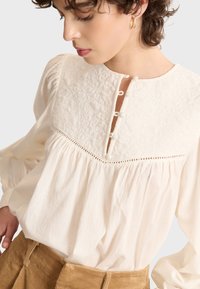 White blouse with textured yoke and front button closure. Features long, puffed sleeves and a high neckline with delicate details.