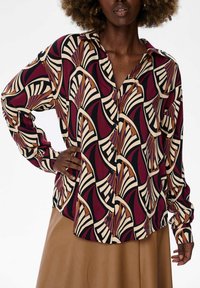 Woman wearing a buttoned shirt with a maroon, cream, black, and brown abstract fan pattern and a tan skirt, hand on hip.