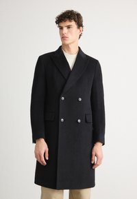 Cappotto nero doppiopetto realizzato in tessuto testurizzato, dotato di due grandi bottoni, rever a punta e tasche laterali, indossato sopra un leggero maglione.