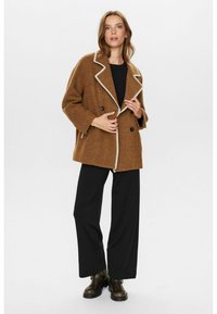 Manteau en mélange de laine marron à double boutonnage avec des coutures blanches contrastantes, coupe oversize et grandes poches avant, associé à un pantalon noir et des bottes.