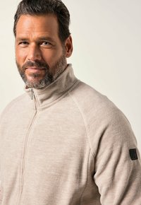 Sudadera de vellón beige con cuello alto, cremallera de media longitud en el frente y costuras en los hombros. Presenta un parche negro de la marca en la manga izquierda.