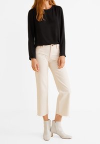 Schwarzes Langarmshirt kombiniert mit cremefarbenen, taillierten Jeans und weißen Ankle Boots. Die Jeans haben rohe Säume und eine enganliegende Silhouette.