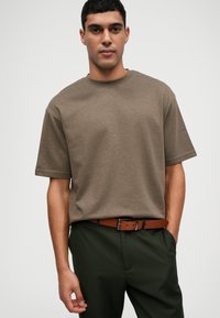 Καφέ βαμβακερό T-shirt με στρογγυλή λαιμόκοψη και κοντά μανίκια, συνδυασμένο με σκούρα πράσινα παντελόνια και καφέ δερμάτινη ζώνη. Λείο υλικό.