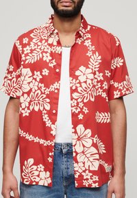 Camisa roja de manga corta con patrones blancos de flores y hojas, diseño con botones, tela suave y etiqueta en el pecho izquierdo. Llevada sobre una camiseta interior blanca.