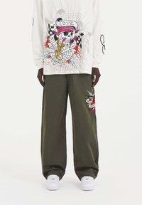 Witte blouse met lange mouwen en grafisch doodshoofd ontwerp; olijfgroene wijde broek met kleurrijke geborduurde accenten. Witte sneakers.