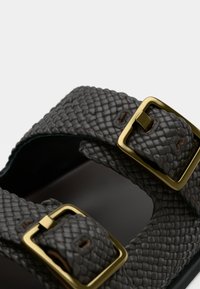 Μαύρο λουρί σανδάλου με υφή, με δύο τετράγωνες αγκράφες από ορείχαλκο και ορατές ραφές σε ανοιχτό φόντο.