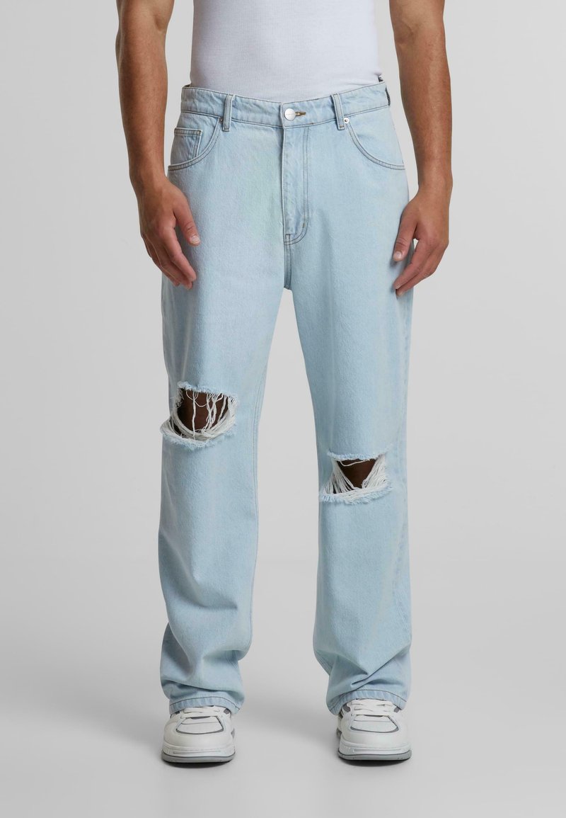 Light blue denim jeans with two large rips at the knees, loose fit, and standard five-pocket design. Styled with white sneakers.