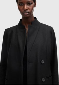 Cappotto in misto lana nero con colletto alto, chiusura a doppio bottone, design elegante e dettagli minimalisti. Tessuto liscio con tasche laterali.