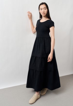 Frau in einem schwarzen, gestuften Maxikleid und beigen Sneakers steht mit einer Hand an einer hellgrauen Wand und schaut zur Seite.