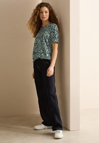 Top de manga corta con estampado en teal y negro combinado con jeans oscuros. El modelo se encuentra de pie contra una pared clara, luciendo un calzado casual.