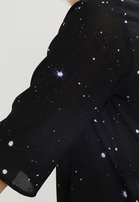 Blusa nera di chiffon con maniche corte, caratterizzata da un motivo stellato di punti bianchi e viola, creando un effetto cosmico. Tessuto traslucido.