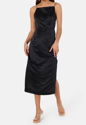 Femme portant une robe midi en satin noire avec fines bretelles et fente latérale, associée à des sandales à talons avec bride transparente sur fond blanc.