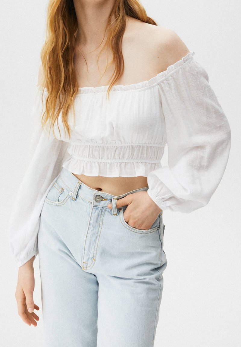 Hvid bluse med bar skulder og elastisk flæse nederst samt lange voluminøse ærmer, kombineret med lyseblå høj talje jeans.