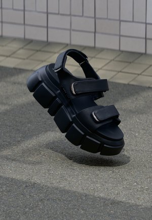 Sort chunky platform-sandal med tyk tekstureret sål og to justerbare velcrolukninger på flise- og asfaltunderlag.