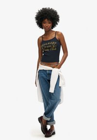 Débardeur court bleu marine avec le logo "Superdry" en jaune, associé à un jean bleu retroussé et des sabots bruns. Chemise blanche nouée à la taille.