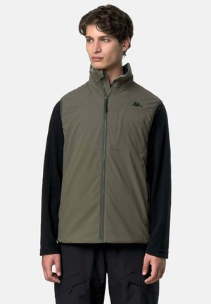 Giovane uomo che indossa un gilet verde oliva senza maniche con cerniera sopra una camicia nera a maniche lunghe, in piedi davanti a uno sfondo bianco uniforme.