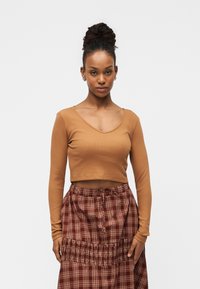 Mladi žena s tamnom kovrčavom kosom u punđi, odjevena u smeđi top dugih rukava i crvenu kariranu suknju na jednostavnoj svijetloj pozadini.