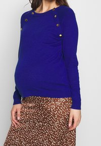 Blå stickad gravidtröja med ribbad detalj och gyllene knappaccenter vid axlarna, i kombination med en brun prickig kjol.