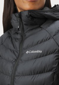 Svart pufferjakke med glidelås, med quiltet tekstur og Columbia-logo på brystet. Høy krage for ekstra varme.