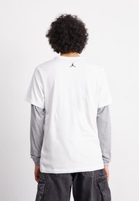 Camiseta blanca de manga corta con mangas largas grises por debajo. Presenta un logo Jumpman negro en la parte posterior, cerca del cuello. Material de algodón.