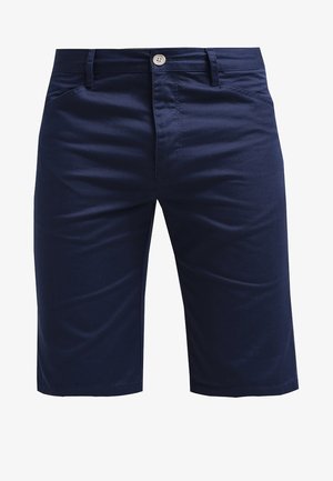 Camisa vaquera abotonada con mangas remangadas sobre una camisa de cuadros, combinada con pantalones cortos azul marino y zapatos blancos sin cordones. Conjunto simple y casual.
