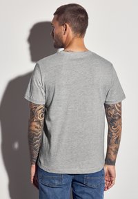 Hellgraues Baumwoll-T-Shirt mit Rundhalsausschnitt und kurzen Ärmeln, das eine glatte Textur und eine lockere Passform aufweist. Sichtbare Tattoos an den Armen.
