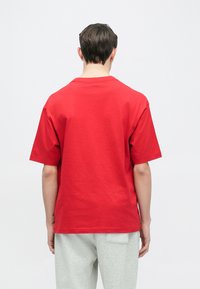 Punainen puuvillainen t-paita, jossa on lyhyet hihat, pyöreä pääntie ja rento istuvuus, yhdistetty harmaisiin nyörillä varustettuihin housuihin. Yksinkertainen, koristelematon muotoilu.