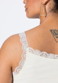Camiseta de marfil con encaje en los tirantes. Textura suave con un diseño ajustado, que muestra un delicado patrón de encaje en el escote.