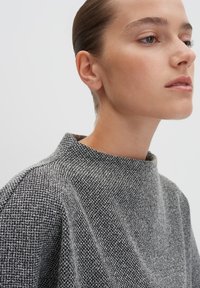 Frau mit nach hinten gekämmtem Haar, die ein hochgeschlossenes, schwarz-weiß strukturiertes Strickoberteil trägt und leicht nach rechts schaut, vor einem schlichten Hintergrund.