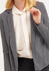 Grå, strukturerad kavaj över en vit blus med slipskrage. Kavajen har en skräddarsydd passform och vågade lapeller, parat med svarta byxor.