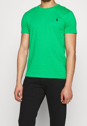 Mann trägt ein leuchtend grünes Kurzarm-T-Shirt mit Rundhalsausschnitt und einem kleinen schwarzen gestickten Logo auf der Brust, kombiniert mit schwarzen Hosen.