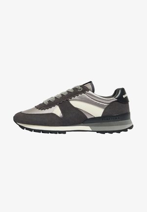 Zapatilla deportiva de ante y tela gris y blanca con cordones en la parte delantera y suela negra, vista desde un lateral sobre fondo blanco.