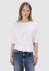 Witte katoenen top met korte mouwen, ronde hals en een ontspannen pasvorm, gecombineerd met blauwe denim jeans. De top heeft een gladde textuur en een iets cropped lengte.