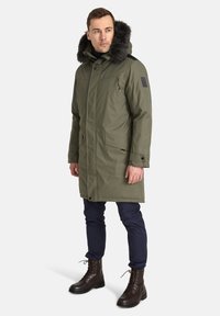 Alyvučių žalios spalvos parka su dirbtinės kailio pamušalu ant kapšiko, priekyje su užtrauktuku, šoninėmis kišenėmis ir juodais akcentais. Dėvima su mėlynais kelnais ir rudais bateliais su raišteliais.