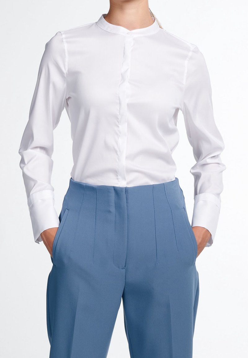 Personne portant une chemise blanche à manches longues boutonnée, rentrée dans un pantalon bleu taille haute sur mesure, les mains dans les poches.