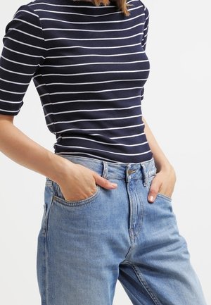 Frau trägt ein marineblau-weiß gestreiftes Hemd, das in eine hoch taillierte hellblaue Jeans gesteckt ist, mit den Händen in den Taschen.