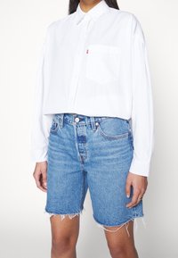 Vit skjorta med knappar och ficka, i kombination med ljusblå denimshorts med fransade kanter. Outfiten har en avslappnad passform och en casual stil.