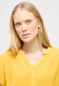 Gul tekstureret bluse med korte ærmer, V-hals og små knapper ned langs fronten. Den har en afslappet pasform og et krøllet stof.