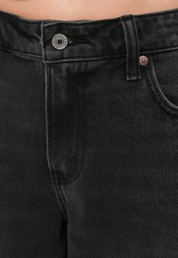 Črne denim hlače imajo ravno kroj, vidne šive, sprednjo zaponko in rahlo teksturo. Ena žepa je delno vidna.