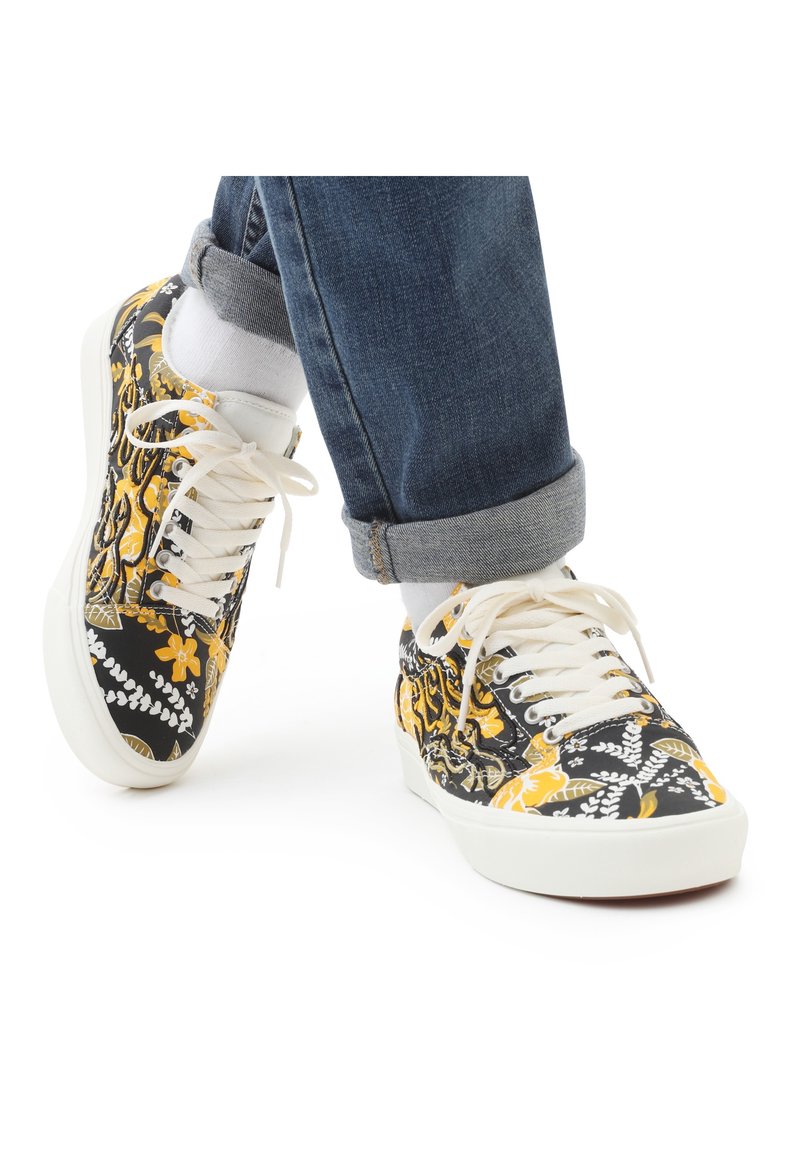 Pieds portant un jean bleu retroussé, des chaussettes blanches et des baskets noires avec des motifs floraux jaunes et blancs sur fond blanc.