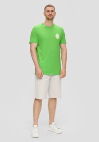 Grünes Baumwoll-T-Shirt mit kurzen Ärmeln und Rundhalsausschnitt, mit einem weißen kreisförmigen Logo auf der linken Seite. Kombiniert mit hellbeigen Shorts.