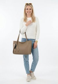 Mujer rubia con suéter blanco, jeans azul claro, zapatillas beige, sosteniendo un bolso grande color topo con etiqueta negra y luciendo reloj y pendientes dorados.