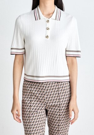 Frau mit weißem, kurzärmligem Stricktop mit gestreiftem Saum, goldenen Knöpfen und gemusterter High-Waist-Hose. Oberkörper sichtbar, Arme seitlich.