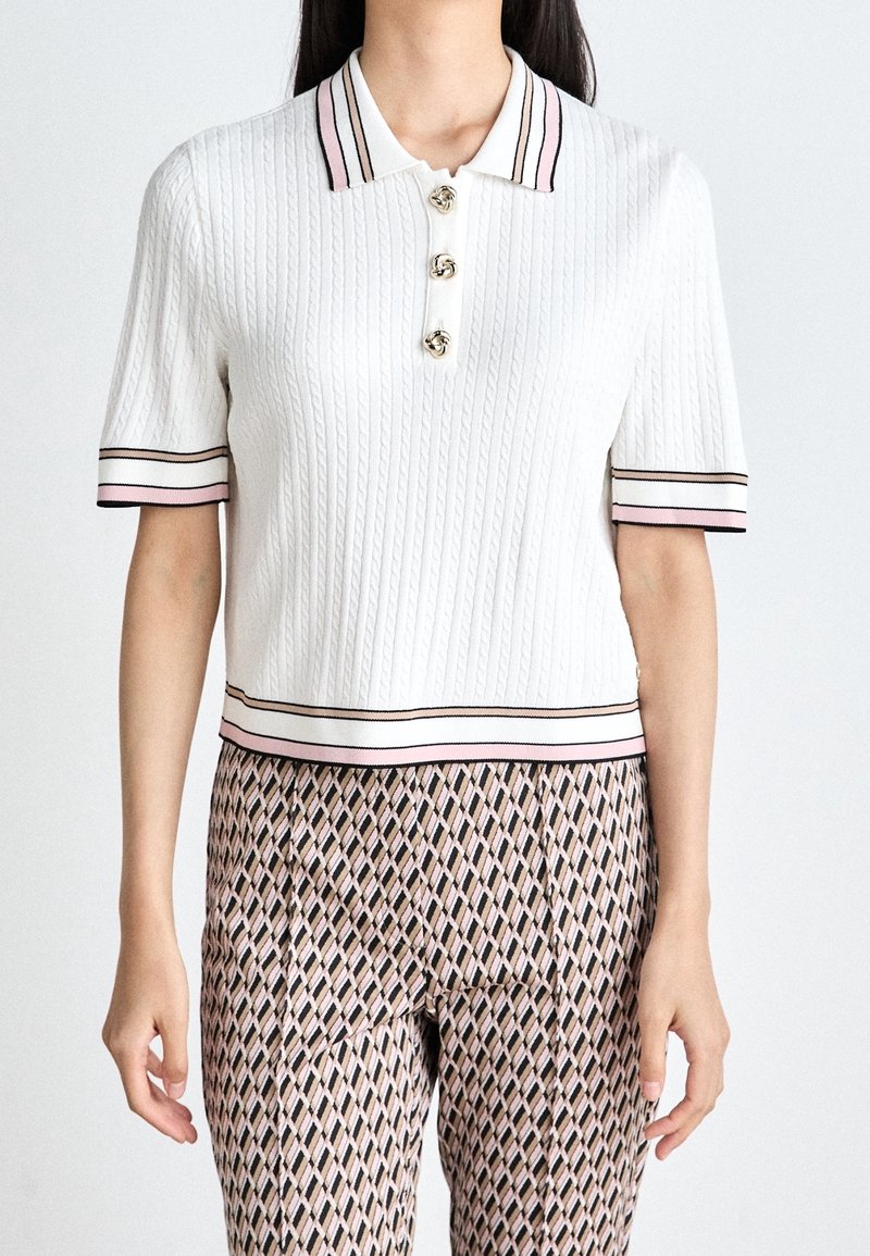 Femme portant un haut en maille blanc à manches courtes avec bordure rayée, boutons dorés et pantalon taille haute à motifs. Buste visible, bras le long du corps.
