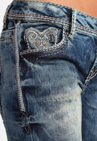 Jeans in denim blu sbiadito con un design a cuore cucito sulla tasca posteriore, accentuato da un filo a contrasto e cuciture decorative.